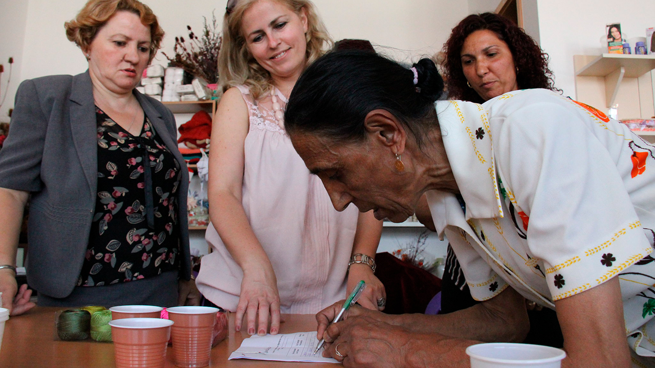 En kvinna från Kosovo skriver på ett papper och anställda vid organisationen Mundesia står i bakgrunden.